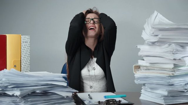 Young female office worker losing temper at work in the office. Stressed and tired manager tearing her hair and screaming in anger. Piles of unfinished paperwork, unfilled documentation and