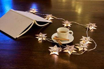 Una taza de café con canela, rodeada de un libro y luces navideñas en forma de estrellas