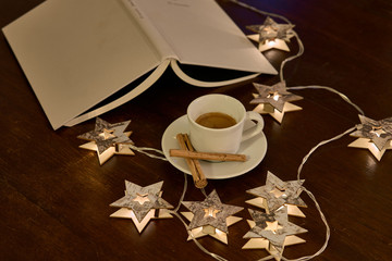 Una taza de café con canela, rodeada de un libro y luces navideñas en forma de estrellas