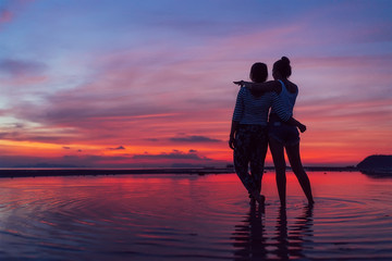Two girlfriends hugging up and enjoying a rose/pink sunset sky on the sea beach on the Samui Island,Thailand. Calm warm countries vacation concept image.