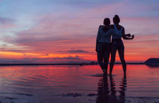Two females intimate hugging up and enjoying a rose/pink sunset sky on the sea beach on the Samui Island,Thailand. Calm warm countries vacation concept image.