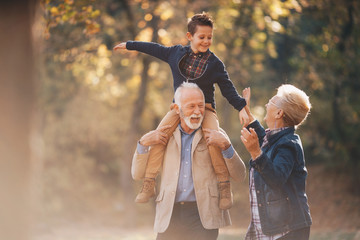 Grandparents having a lovely autumn day with their grandson in nature. The grandfather is carying his grandson on his shoulders while the grandmother holds the child's hands. - obrazy, fototapety, plakaty