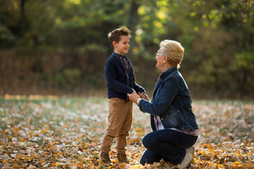 Happy grandmother sitting on one knee and holding hands with her little grandson in the park in autumn.