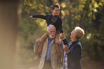 Grandson on his grandfather shoulders and smiling and holding hands with his grandmother during the walk through park.