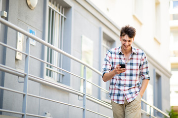 Fototapeta premium smiling young man walking and looking at mobile phone outside