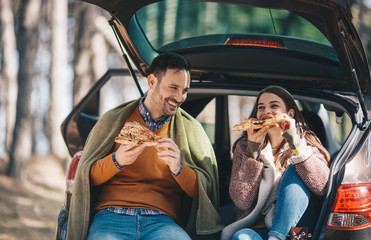 Young cheerful Caucasian couple making a stop and eating pizza while they are sitting on open cat...