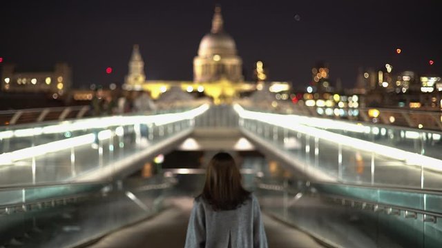 Tracking shot of a tourist woman walking on London Millennium Bridge with St Paul Cathedral view lights at night