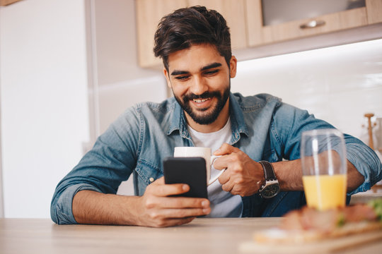 A man using his smartphone and holding a mug of coffee in the kitchen. There is a glass of orange juice as well.