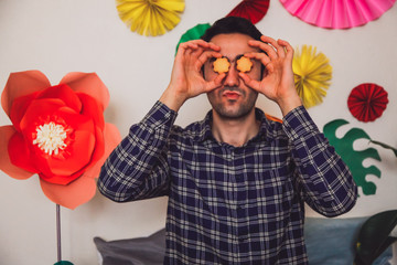 A brunette man holding two flower shaped cookies next to his eyes with flowers in the background....