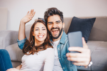 Beautiful young Caucasian couple smiling and taking a selfie while they are sitting by the couch and waiting for a tv show to start in their living room. - obrazy, fototapety, plakaty