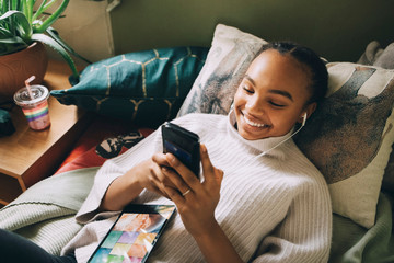 High angle view of smiling teenage girl listening music while using mobile phone on bed at home