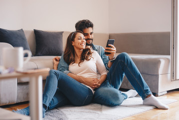 Obraz premium Cute Caucasian newly weds watching photos on smart-phone while they are embracing and relaxing on the floor by the couch.