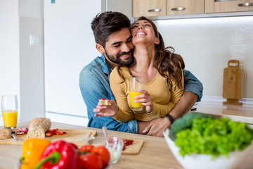 Young handsome Caucasian man hugging his beautiful cheerful girlfriend with love while they are having breakfast in the kitchen.