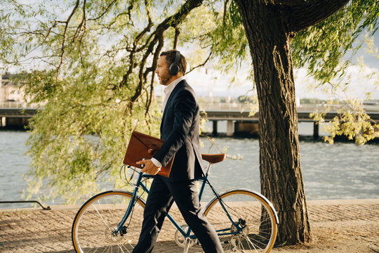 Side view of businessman walking with bicycle on footpath while listening music through headphones
