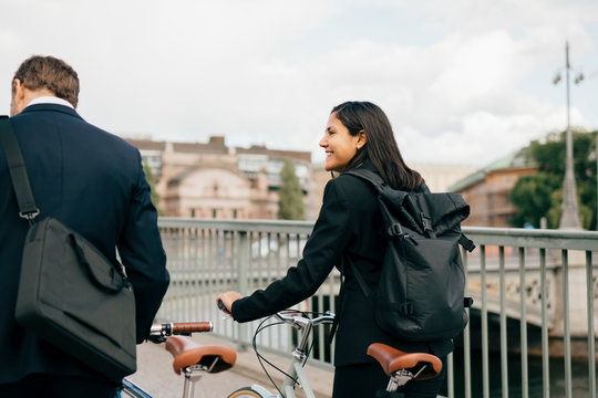 Smiling businesswoman looking at businessman while walking with bicycles on bridge in city - Powered by Adobe