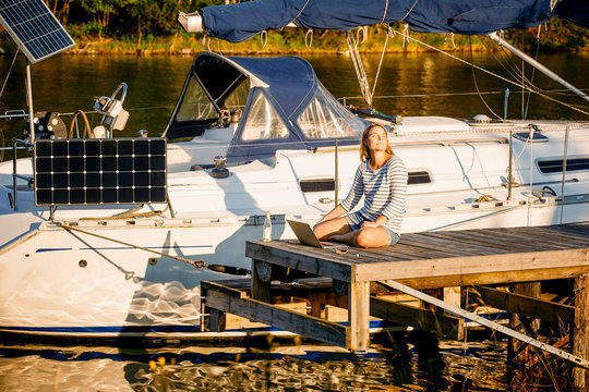 Thoughtful woman with laptop sitting on pier by moored yacht at sunny lakeside