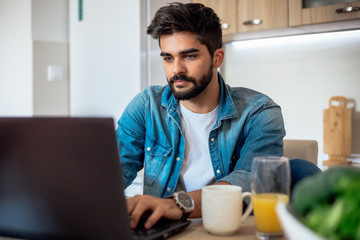 Attractive young bearded Caucasian freelancer working on his laptop while sitting in the kitchen. - obrazy, fototapety, plakaty