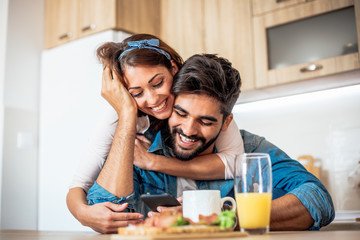 Young Caucasian couple in love, embracing and watching photographs from their vacation on the smart-phone in the kitchen.