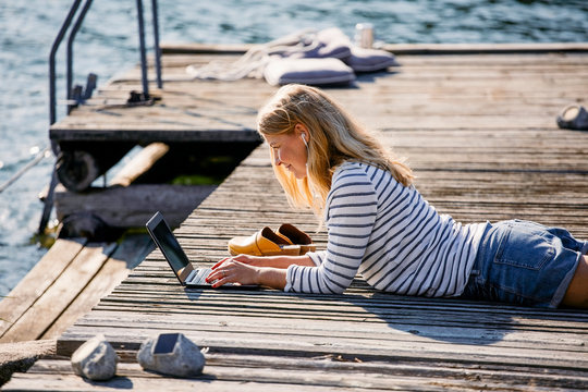 Mid adult female typing on laptop while lying over wooden pier during summer vacation