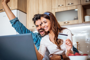 Young beautiful Caucasian couple happy and excited with successful purchase they just made on their laptop while they are sitting in the kitchen.