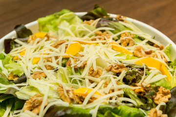 Closeup of delicious salad of lettuce, watercress, arugula, mango, walnuts and shredded cheese on white ceramic platter on wooden table. Healthy and nutritious food. Selective focus.