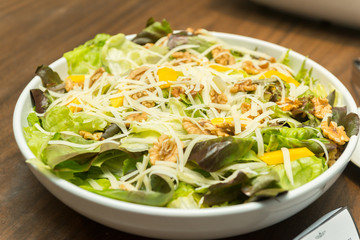 Closeup of delicious salad of lettuce, watercress, arugula, mango, walnuts and shredded cheese on white ceramic platter on wooden table. Healthy and nutritious food. Selective focus.