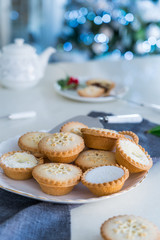 Obraz na płótnie Canvas Traditional english festive pastry mince pies on served for tea time table with lightened christmas tree on background. Cozy home mood. Vertical card. Close up, selective focus. Copy space.