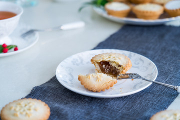 Tea time table setting with broken Mince Pie with filling on white plate. Traditional english festive pastry. Cozy home mood. Close up, selective focus. Copy space.