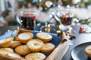 Warm cosy composition of traditional english festive pastry mince pies in wooden tray with blurred background of mulled wine drinks, lights garland and christmas tree on background. Close up. © okrasiuk