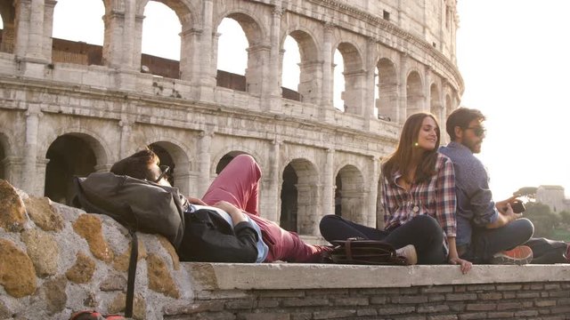 Three young friends tourists sitting in front of colosseum in rome at sunset taking selfies with smartphone with backpacks sunglasses happy beautiful girl long hair slow motion