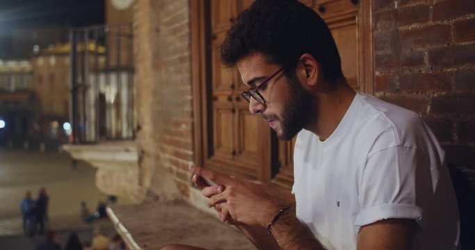 Portrait of latin handsome boy using a smarthone sitting outdoors. Closeup sociable young man with mobile phone against beautiful architecture building. Italian guy. Siena, Tuscany