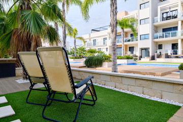 Two empty lounge chairs on artificial lawn grass inside of personal area backyard residential summer villa, view to modern homes with swimming pool, luxury, summer holidays lifestyle concept
