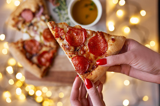 Christmas and New Year atmosphere.Womans hand takes slice of Italian pizza with melting tomato,pepperoni and cheese on a white marbel cutting board.Background with lights in bokeh and selective focus