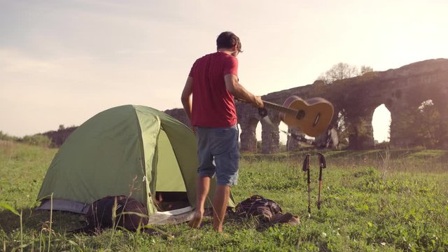 Young man backpacker outside camping tent picks up guitar and walks on grass in front of roman aqueduct arches in parco degli acquedotti park ruins in rome at sunset sticks in ground slow motion