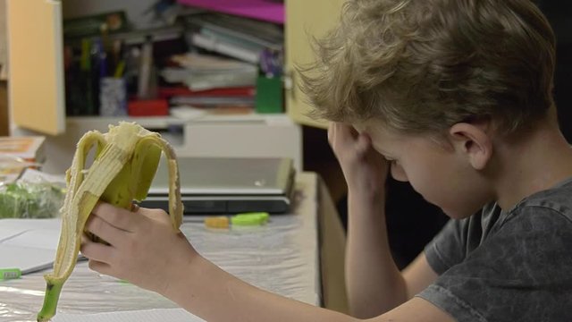 A boy with a banana in his hand draws a pen on a piece of paper. A keen boy sitting at home at a table draws on a piece of paper with a blue pen holding a banana in hand and deliciously bites off it.