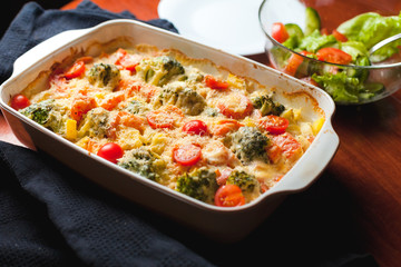 Ready-made baked vegetables in a white ceramic bowl with grated cheese and tomatoes on black towel on table . Natural style