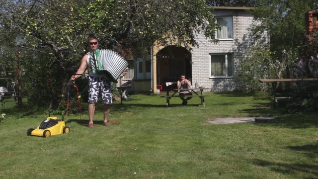 Funny man is mowing lawn by lawnmower, playing harmonica.   Funny, humor, humorous.