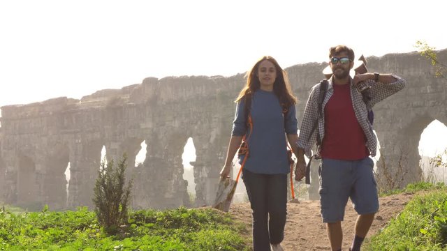 Young lovely couple backpackers tourists walking holding hands toward roman aqueduct arches in parco degli acquedotti park ruins in rome on romantic misty sunrise with guitar and sleeping bag slow