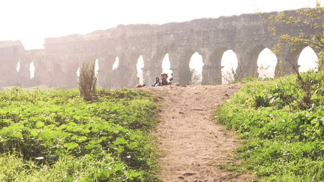 Young lovely couple backpackers tourists walking holding hands toward roman aqueduct arches in parco degli acquedotti park ruins in rome on romantic misty sunrise with guitar and sleeping bag slow
