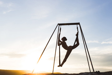 Slim woman practicing fly dance acrobatic yoga poses in hammock outdoors at sunset. Sports and wellbeing concept. Healthy lifestyle.