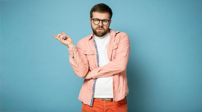 Pompous bearded man with glasses, shows a hand gesture to the side and looks at the camera. Isolated on a blue background.