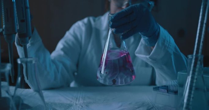 Female research scientist mixing up a conical flask.Blue lighting in a dark lab room.Medium, dolly out, slow motion, shot with BMPCC 4K.Concept: chemistry,science