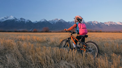 Fototapeta premium One caucasian children walk with bike in wheat field. Little girl walking black orange cycle on background of beautiful snowy mountains. Biker stand with backpack and helmet. Mountain bike hardtail.