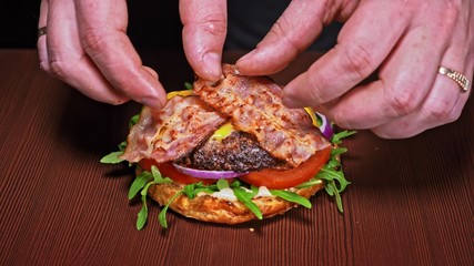 Craft burger is cooking on black background. Consist: sauce, arugula, tomato, red onion, bacon, red currant sauce, ricotta cottage fresh cheese, air bun and marble meat beef. Not made ideal. Looks