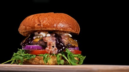Craft burger is cooking on black background. Consist: sauce, arugula, tomato, red onion, bacon, red currant sauce, ricotta cottage fresh cheese, air bun and marble meat beef. Not made ideal. Looks