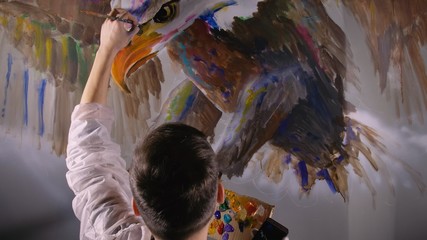 Fototapeta premium Artist designer draws an eagle on wall. Craftsman decorator paints picture with acrylic oil color looking at sketch in phone. Painter painter dressed in paint coat. Indoor. Dark magic cinematic look.