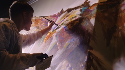 Artist designer draws an eagle on wall. Craftsman decorator paints picture with acrylic oil color looking at sketch in phone. Painter painter dressed in paint coat. Indoor. Dark magic cinematic look.