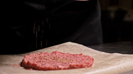 Cook makes meat medallions for burgers. Chef in black food gloves makes cutlet. Cutlets are leveled in steel ring in an even medallion. On top of pouring spices pepper and salt.