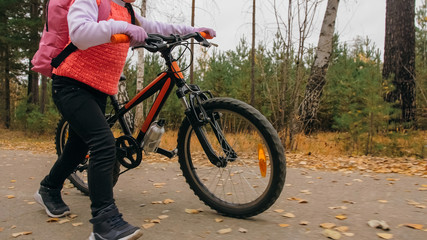 One caucasian children walk with bike in autumn park. Little girl walking black orange cycle in forest. Kid goes do bicycle sports. Biker motion ride with backpack and helmet. Mountain bike hardtail.