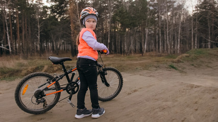 One caucasian children rides bike road track in dirt park. Girl riding black orange cycle in racetrack. Kid goes do bicycle sports. Biker motion ride with backpack and helmet. Mountain bike hardtail.